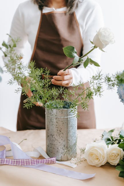 A person dressed in a white top and brown apron arranging a simple floral display with a single white rose and green foliage in a rustic, silver metal vase. The arrangement features delicate white ros
