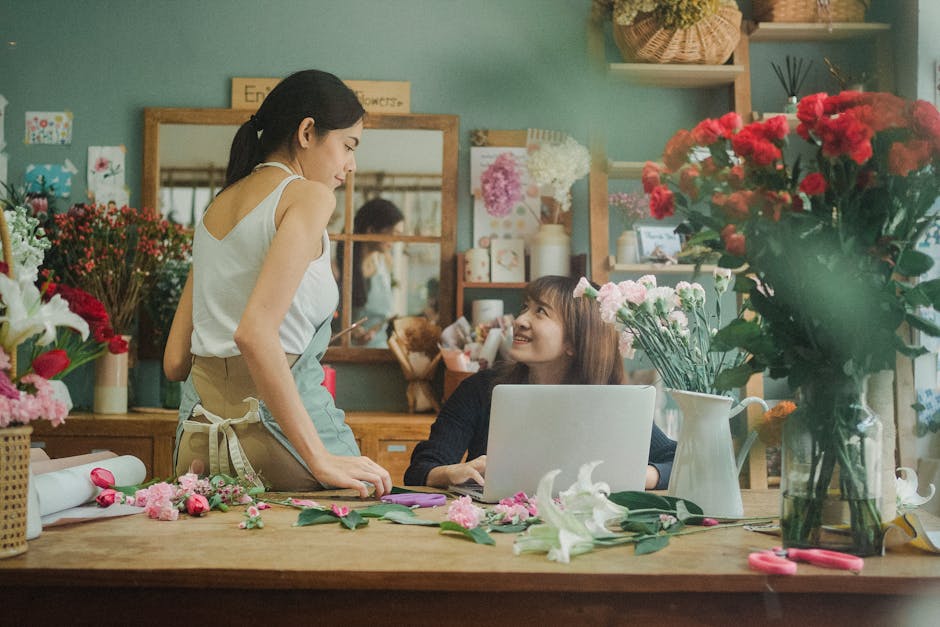 A woman and a florist assistant are engaged in a floral arrangement within a flower shop. The woman, standing on the left, is dressed in a white sleeveless top and a beige apron, leaning slightly forw