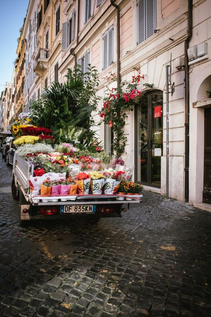 A delivery truck parked on a cobblestone street in front of a European-style building with light-colored stone walls and blue window shutters. The truck's bed is filled with an assortment of fresh flo