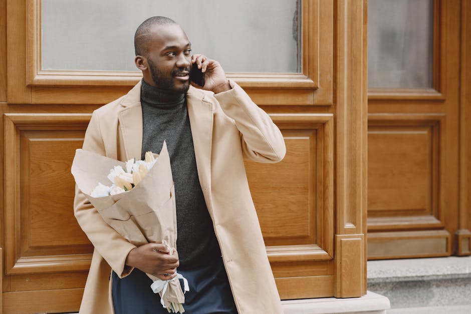 A man with a neatly trimmed beard and short hair, dressed in a beige coat over a dark grey turtleneck, stands outdoors in front of a wooden-paneled building. He holds a bouquet of fresh, predominantly